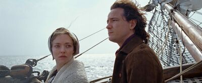 A woman wearing 1800s clothing and a man stand together on a windy ship in the ocean