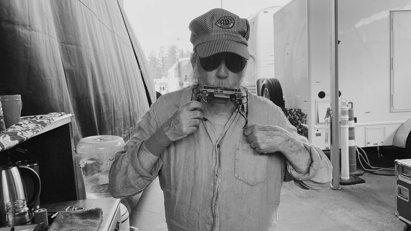 Neil Young wears a base ball cap and sunglasses and plays the harmonica.