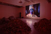 A large video screen in a gallery shows a character wearing blue face paint and an elaborate costume. 