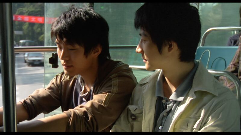 Two men sit on a bus. One of them looks at the other one smiling,.