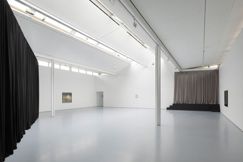 Photograph shows an installation in a brightly lit gallery with white walls and grey floor. There are large black pleated curtains hanging in the space, nearly to the ceiling. These are hung in broad sweeping diagonals across the large floorspace. On the right of the photograph, at the back of gallery two can be seen a beige curtain and a low black platform with small steps leading to it from either side, as though a stage. In the far corner, to the left hand side, we can see a framed photograph on a white wall in the distance. 