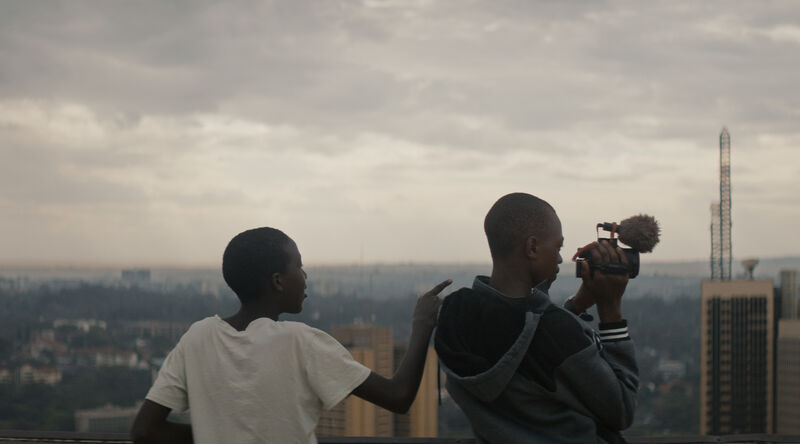 Two boys stand on a roof top looking out at a city, one of them is holding a camera.