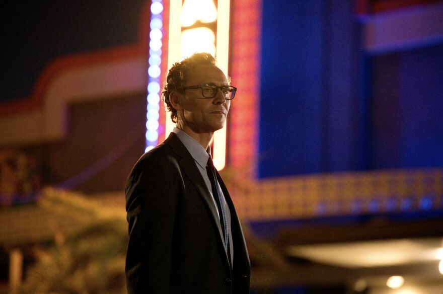 A man wearing a suit and glasses stands outside of a theatre with bright lights.