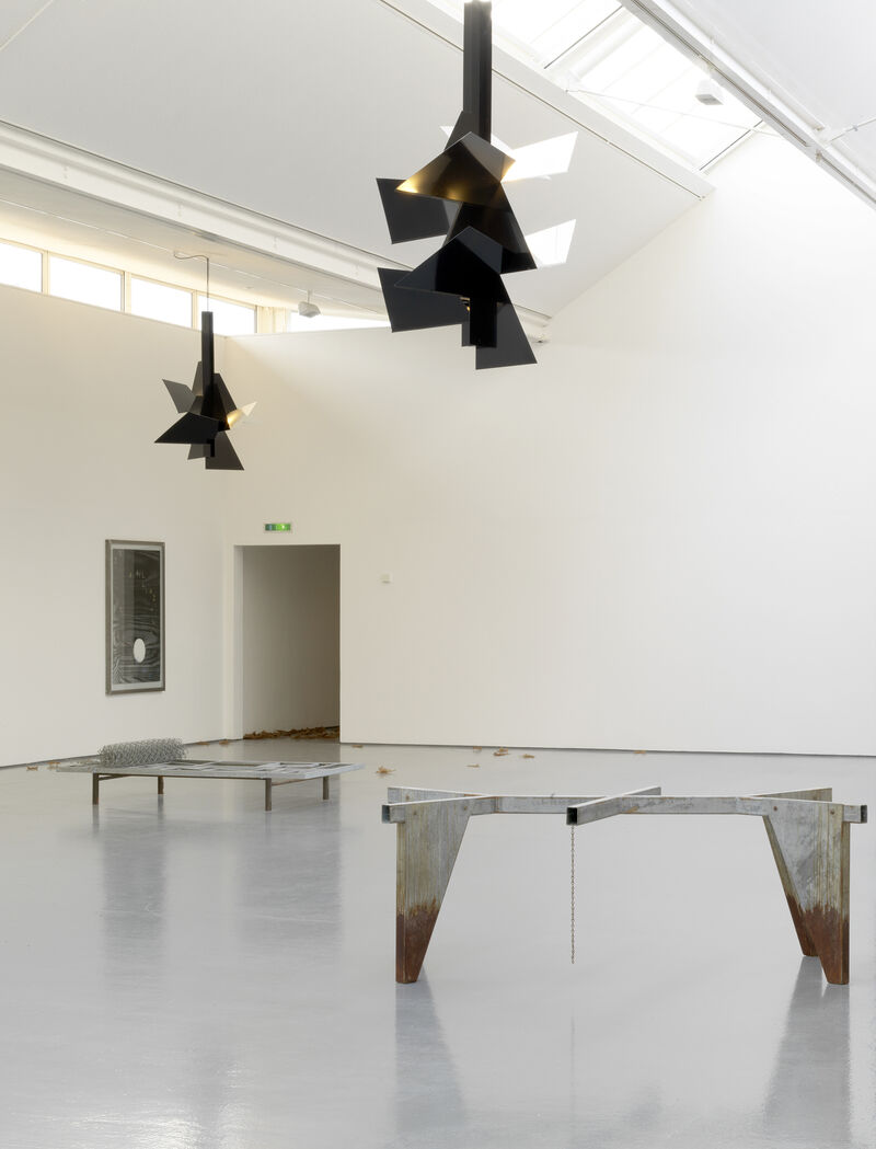 From Martin Boyce's exhibition at DCA. Black, triangular light structures hang from the ceiling. There are metal sculptures on the floor. The sculptures are sharp and industrial looking.