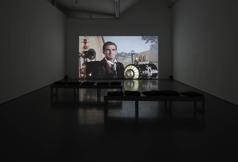 A projector in DCA Galleries from Thomas & Craighead's exhibition shows a vintage film of a man sitting at a futuristic looking device.