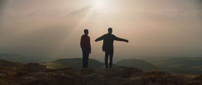 Two boys stand on a hill together with their arms outstretched, the sun obscuring them.