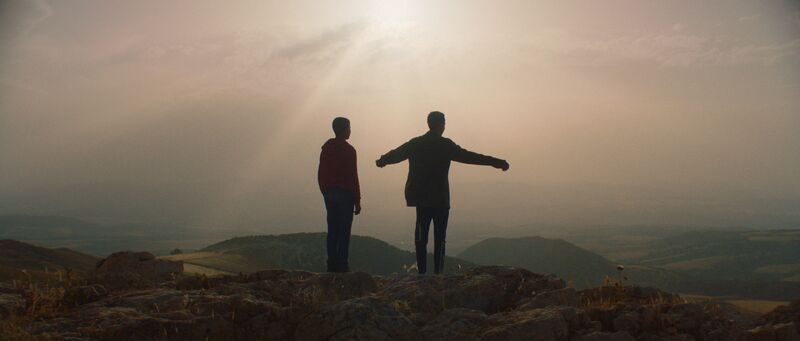 Two boys stand on a hill together with their arms outstretched, the sun obscuring them.