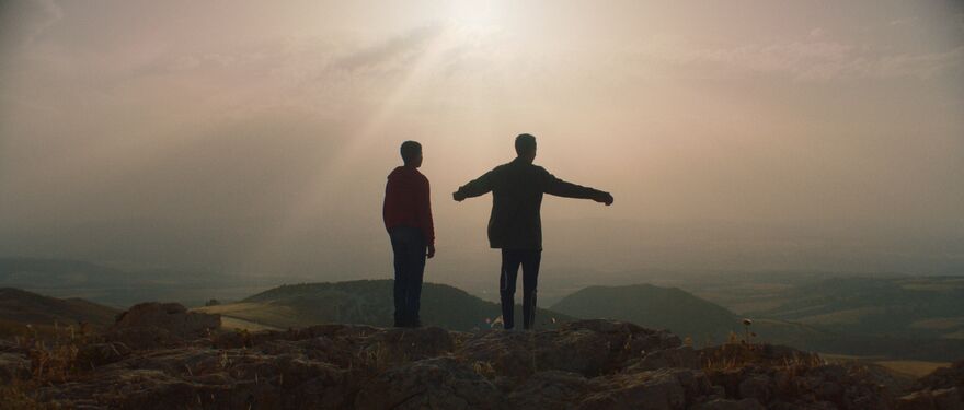 Two boys stand on a hill together with their arms outstretched, the sun obscuring them.