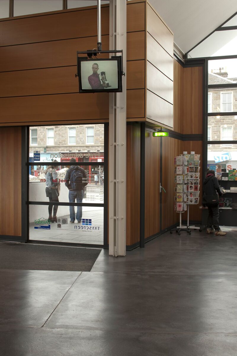 An image from Nikolaj Bendix Skyum Larsen' exhibition at DCA. DCA reception is pictured. There is a televisions screen hanging from the ceiling which has an image on it of a man wearing a pink shirt holding a cat.