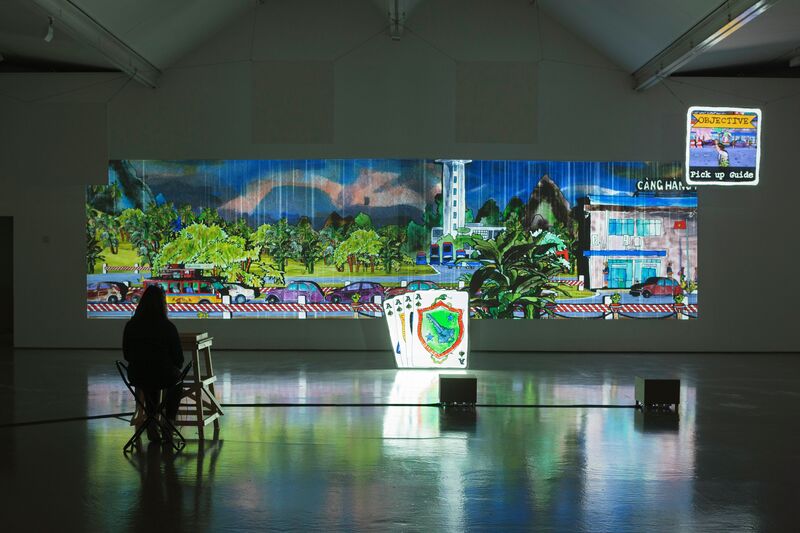 Eddo Stern's exhibition in DCA Galleries. Two projectors show a mixed-media image of cars driving on the road, with a grey rainy background and trees. An illuminated sculpture of a deck of cards is in front of the projector.