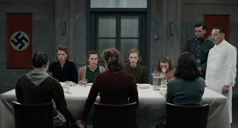 A grouo of 1940s women sit at a dining table in a grey room, with a Nazi flag behind them and two men watching them