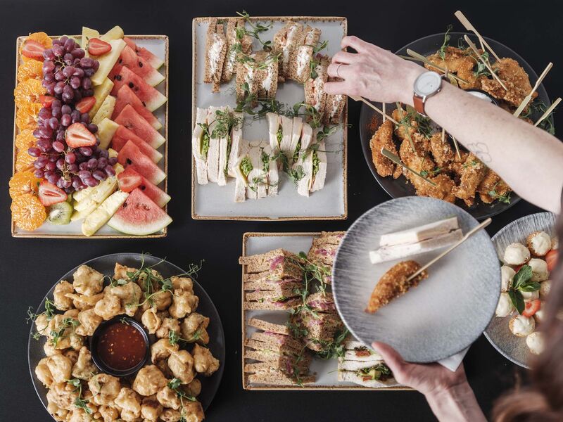 A catering spread including a fruit platter, a sandwich platter, chicken gougjans on sticks, tempura vegetables. A hand reaches over to pick up a sandwich with their half full plate over