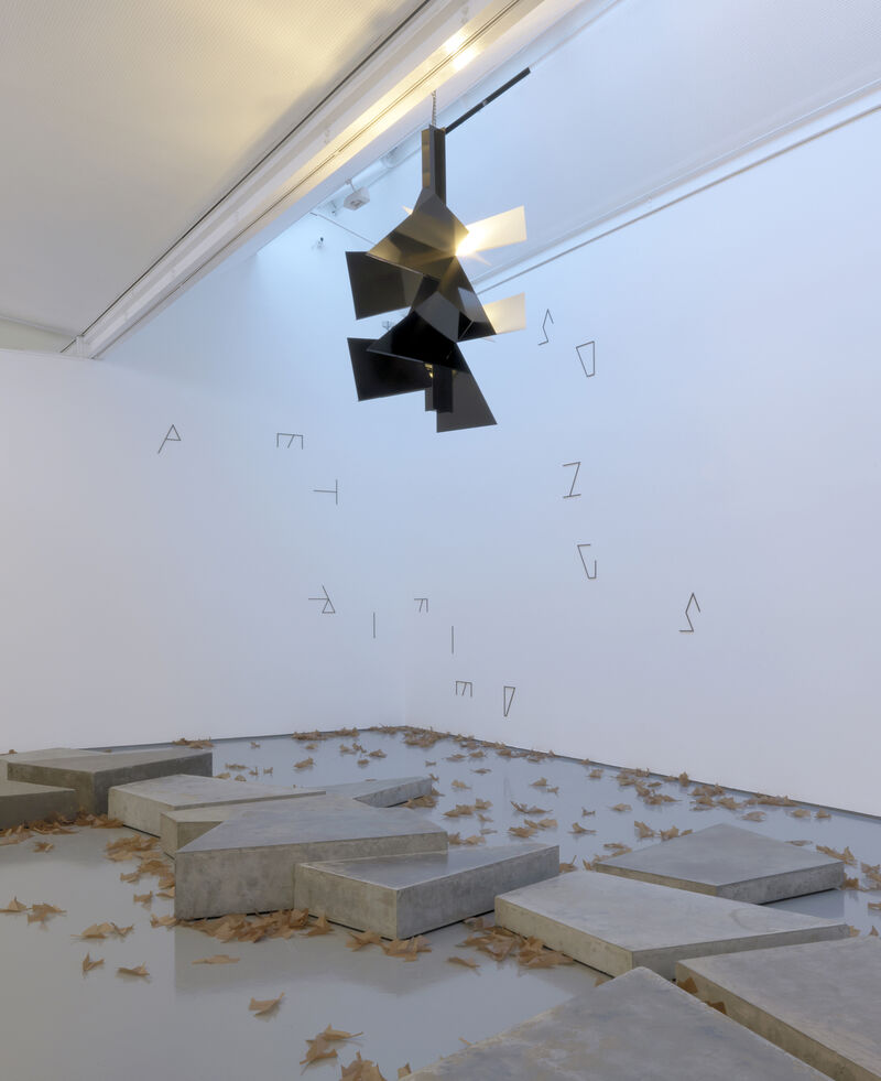 From Martin Boyce's exhibition at DCA. A black, angular and triangular light structure hangs from the ceiling. There are angular letters on the white gallery walls. On the floor, there are large, grey concrete blocks.