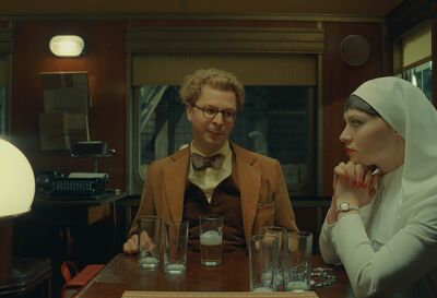 A man in a camel suit, glasses and a bow tie smiles at a nun dressed in white.