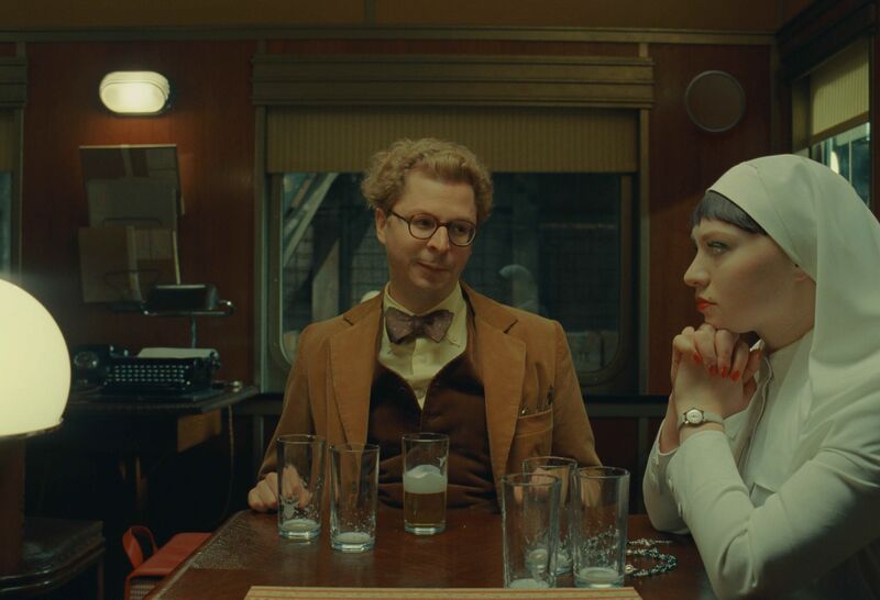 A man in a camel suit, glasses and a bow tie smiles at a nun dressed in white.