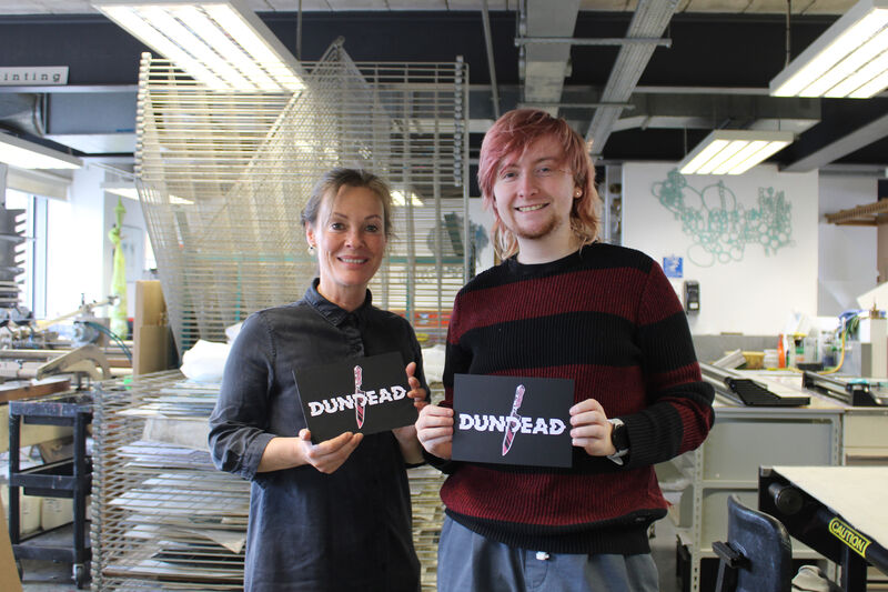 Print Studio Technichian, Claire McVinnie, and Dundead illustrator, Liam Doyle in DCA Print Studio holding Dundead 2024 screen prints
