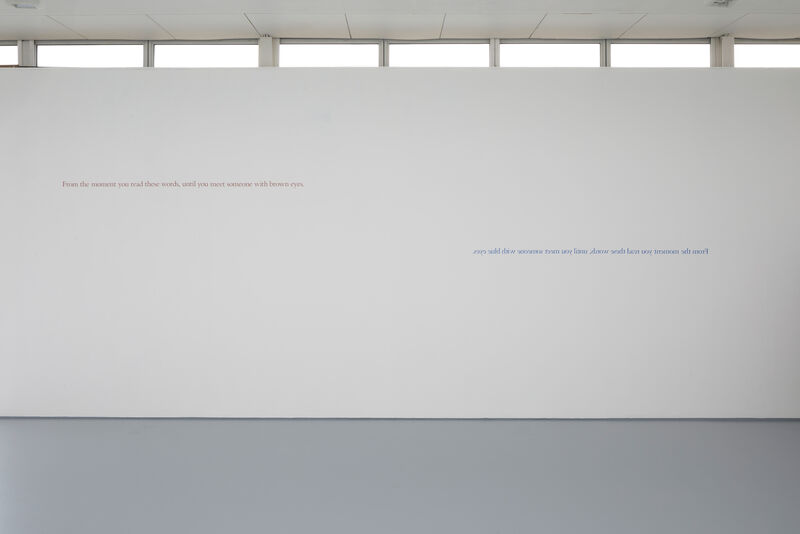 This image shows a text artwork in vinyl on the gallery walls. Higher up on the left hand side, brown vinyl reads: 