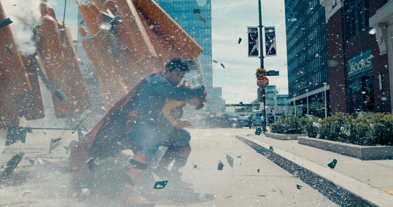 Superman stands on a city street shielding his face from an explosion.