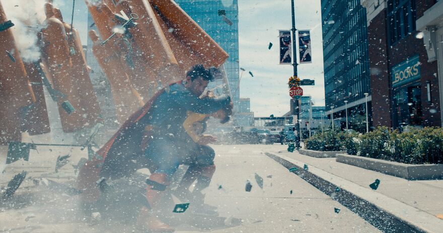 Superman stands on a city street shielding his face from an explosion.