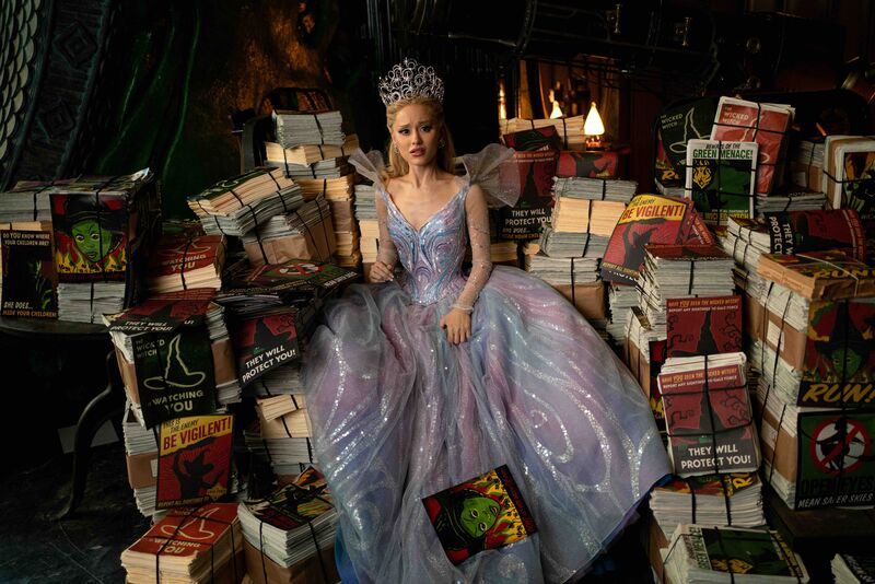 A woman with a large tiara and a princess dress on sits nexts to stacks of posters