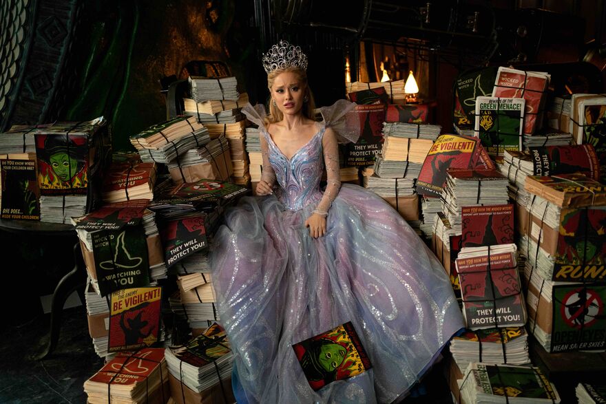 A woman with a large tiara and a princess dress on sits nexts to stacks of posters