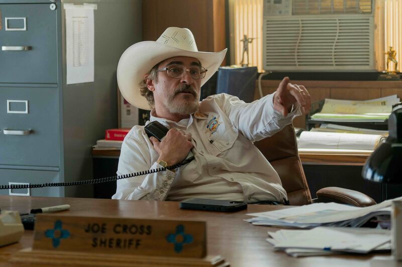 A man in a button-up shirt and a cowboy hat sits at a desk, holding a phone and pointing.