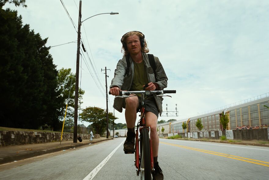 A man with long, shaggy hair cycle a bicycle down a road while wearing headphones
