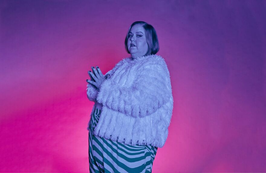 A person wearing a white fluffy cardigan stands against a pink and purple background.