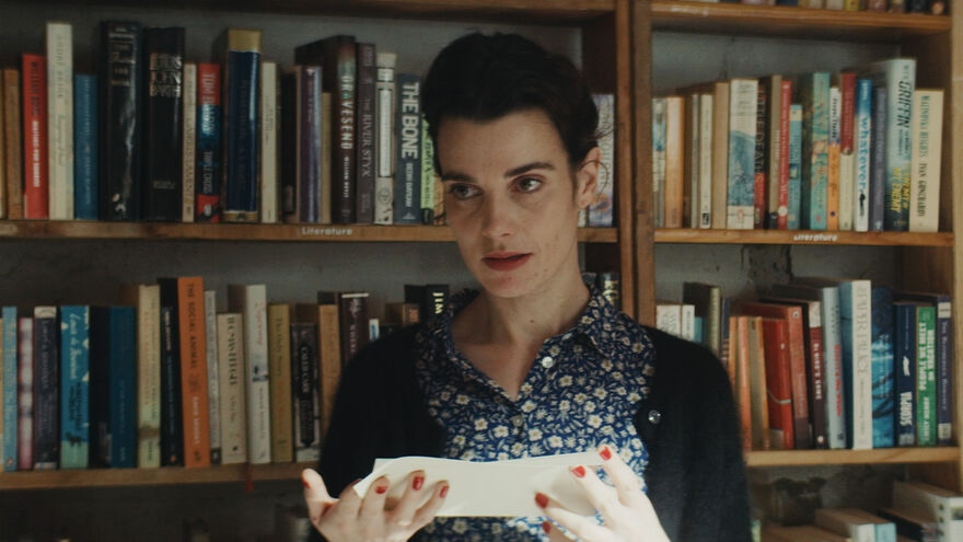 A woman stands in front of a book shelf looking at a note.