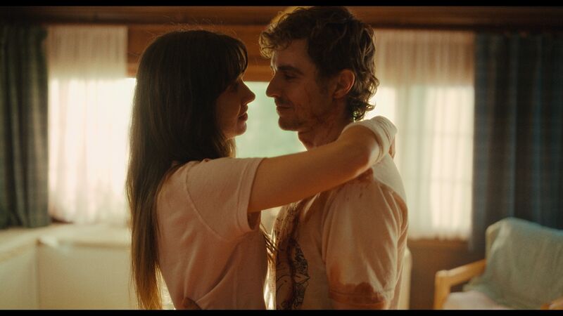 A man and woman embrace. The man has blood on his shirt and the woman has a bandage on her arm. They both smile.