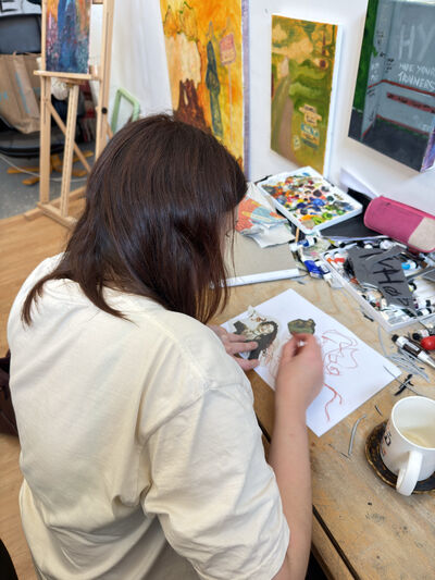 A person with long brown hair drawing at a messy art desk