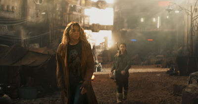 Two people walk through a large industrial-looking environment filled with machinery, structures and atmospheric light filtering through the background.