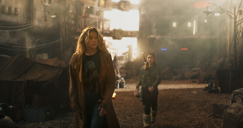 Two people walk through a large industrial-looking environment filled with machinery, structures and atmospheric light filtering through the background.