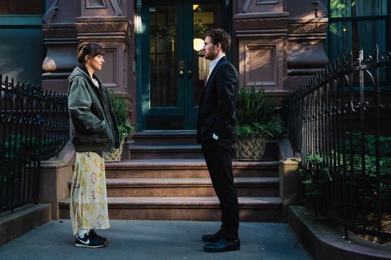 A woman in a big hoodie talks to a man in a suit on the steps of an apartment