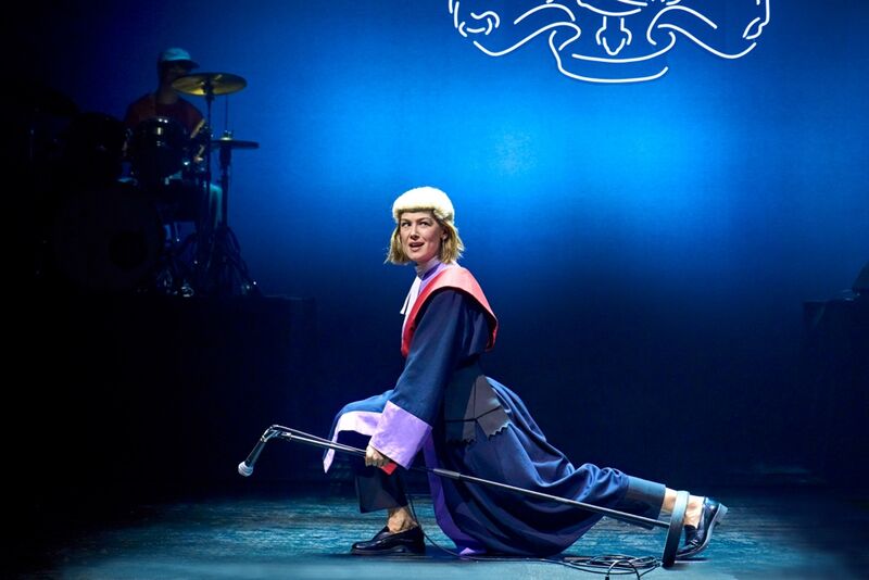 A woman wearing a judge's outfit strikes a pose on stage while holding a microphone