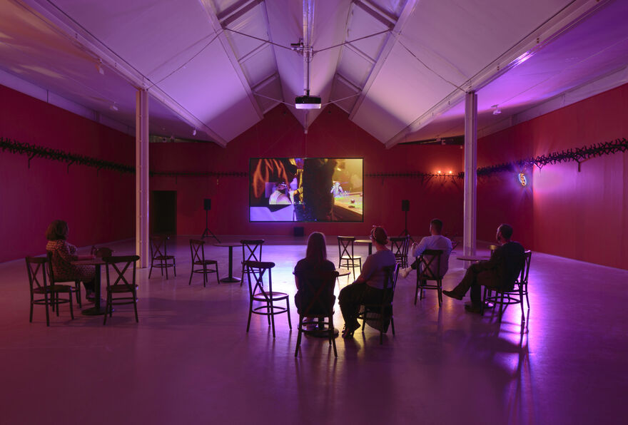 A wide shot photograph of gallery two, showing the gallery with red walls and cabaret-style seating with small tables in the floorspace. A few people sit and face a screen with a projected film. 