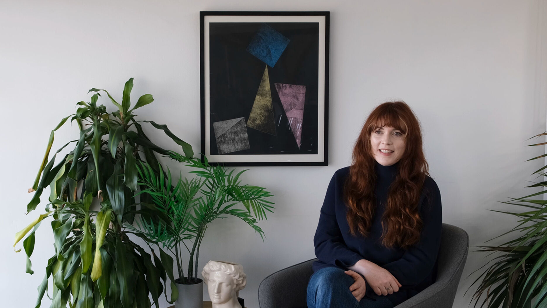 A girl with long red hair sits smiling at the camera. There is a plant to her left and a geometric picture on the wall