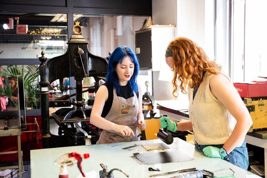 Two people in the Print Studio. One is rolling ink using a roller and the othe is providing instruction