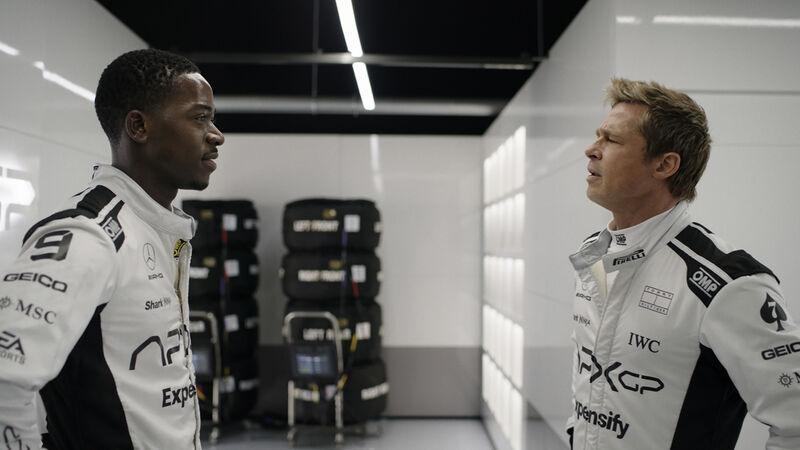 Two men in formula racing car uniforms look at each other.