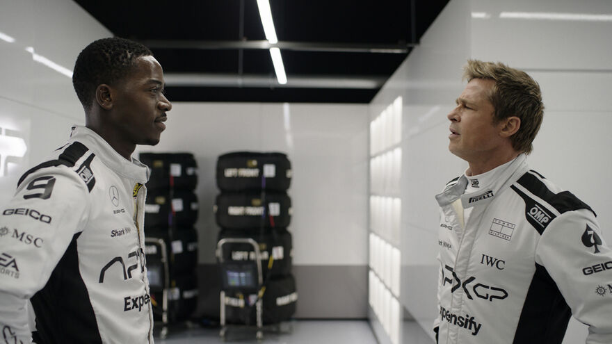 Two men in formula racing car uniforms look at each other.
