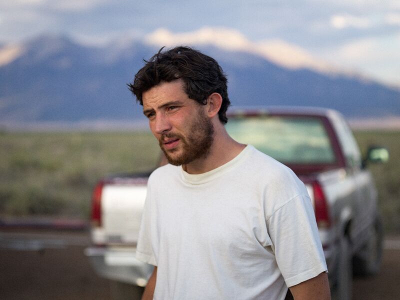 Josh O'Connor wearing a white t shirt with a silver pick up truck and mountains in the background