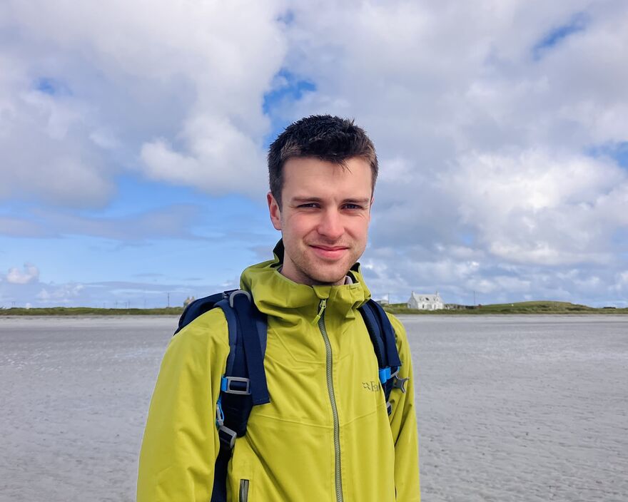 Photo of Sam Andres on a beach wearing a green jacket and navy backpack