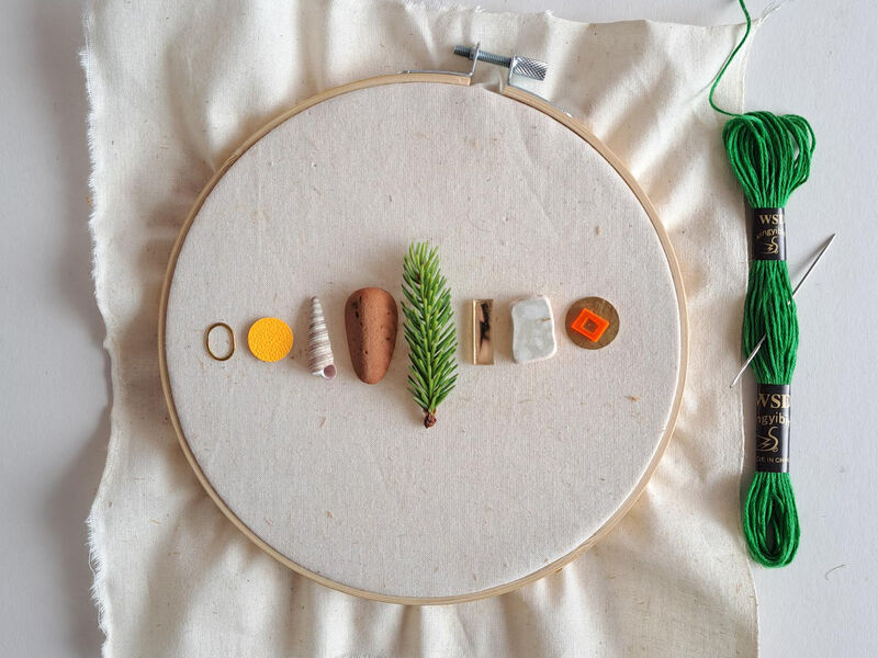 Selection of objects sitting in the middle of an embroidery hoop covered with calico fabric. Green embroidery thread and a needle sit to the righ hand side