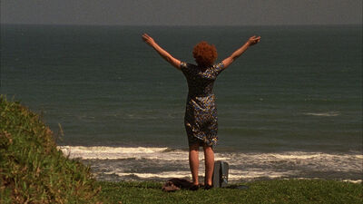 A woman with ginger hair stands at the shore with her arms in the air.