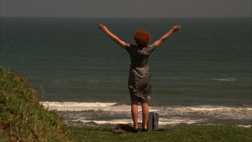 A woman with ginger hair stands at the shore with her arms in the air.