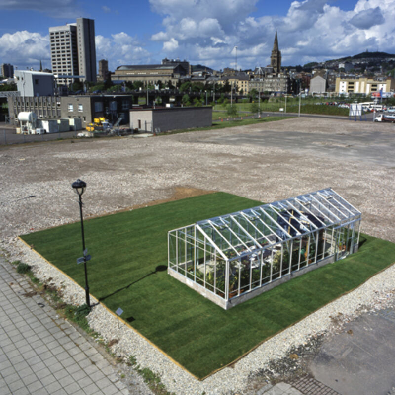From Our Surroundings at DCA. A square of grass sits in an empty plot of land in Dundee. On the grass square, there is a large glass conservatory.