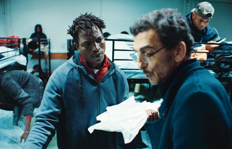A man holds a parcel while speaking to another man in a large warehouse with beds in it
