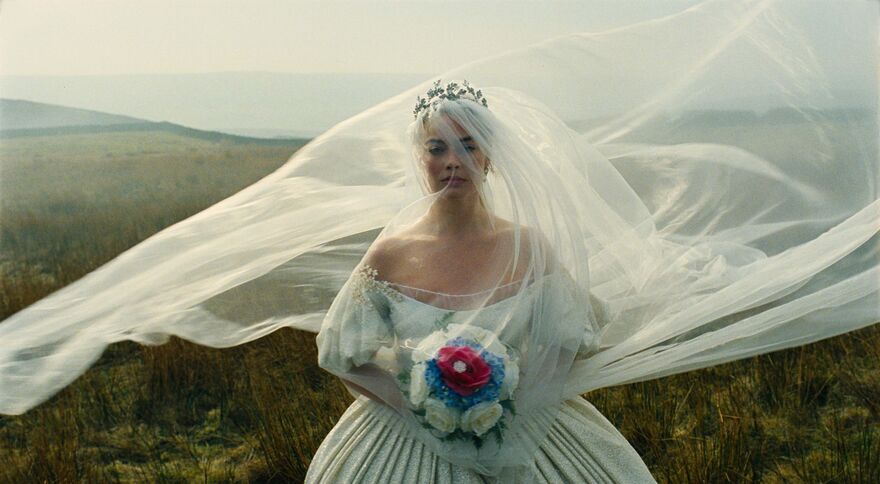 A serious woman in a wedding dress with a veil blowing in the wind stands in a field