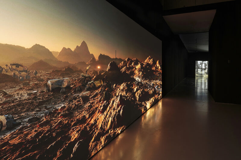 Image shows a film projection, looking along the wall at a sharp angle towards a door. The film still shows a rocky landscape, like the surface of mars. The gallery has a warm glow from the light of the projection.