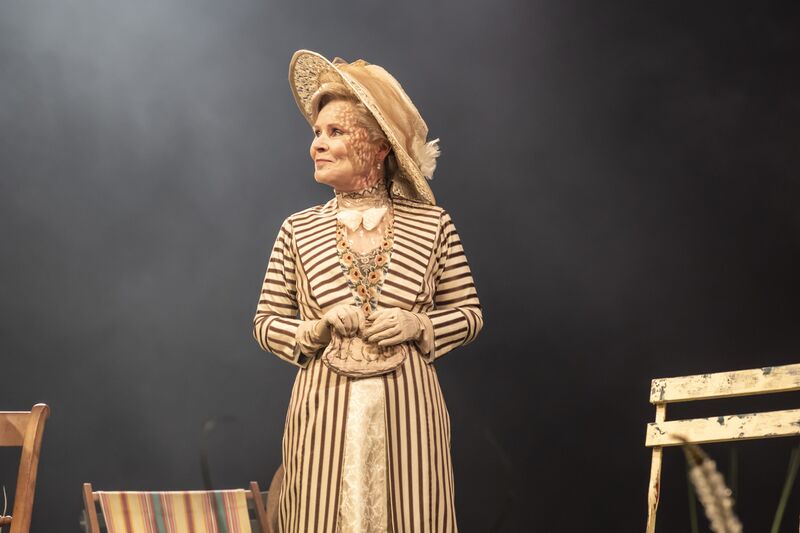 A woman in a large straw hat and a stripy 1900s dress stands on stage.